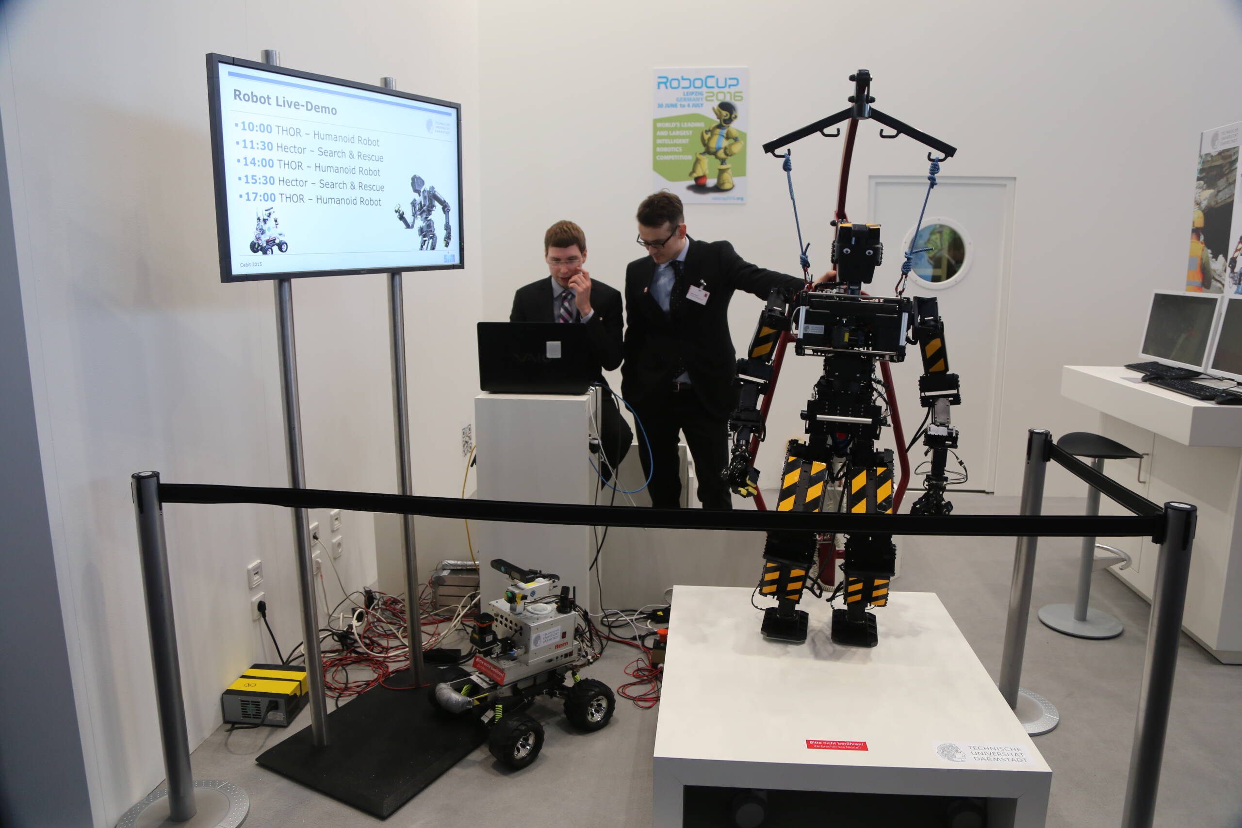 A THOR humanoid robot at a live demo event alongside a Hector search-and-rescue platform. The rigging required to keep the humanoid upright and the controlled demo environment illustrate the gap between exhibition performance and operational deployment.